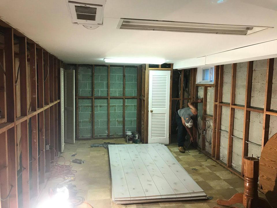Room under renovation with exposed wall studs, stacked drywall sheets on the floor, and a person working near the back wall.